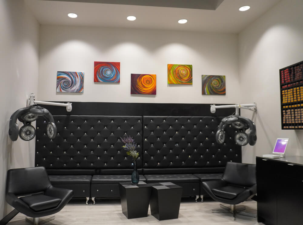 - Studio Wish Salon Spa, OH A modern salon waiting area with black leather seating, two dryers, geometric tables, colorful abstract art on the wall, and a small flower arrangement. - Studio Wish Salon Spa, OH