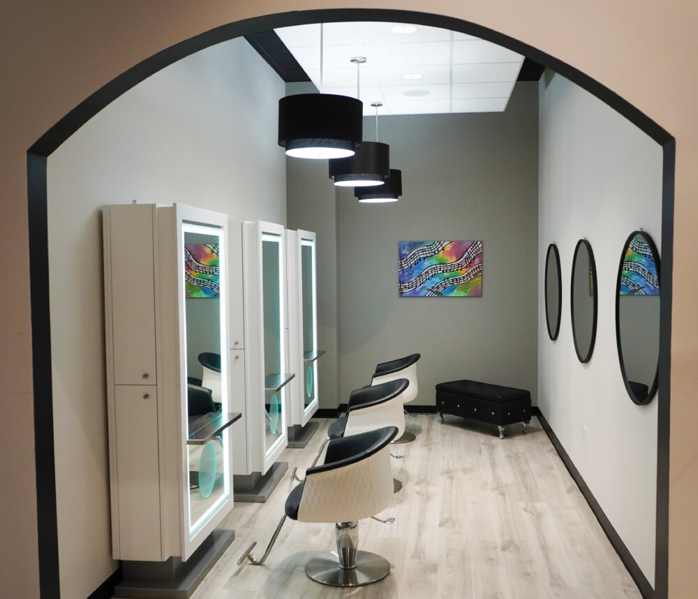 - Studio Wish Salon Spa in Twinsburg, OH A modern luxury salon spa room with three styling chairs facing mirrors, white cabinets, round black mirrors on the right wall, and colorful artwork on the gray back wall—perfect for Aveda-inspired skincare facials. - Studio Wish Salon Spa in Twinsburg, OH