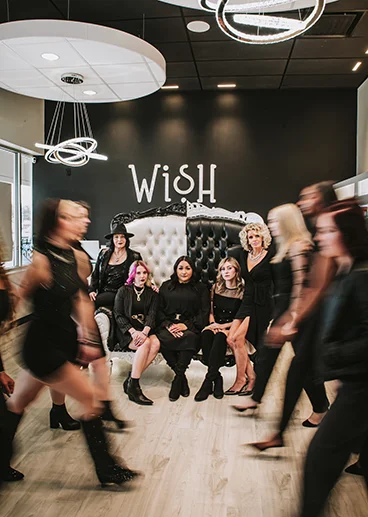 - Studio Wish Salon Spa in Twinsburg, OH A group of women poses on and around a decorative couch under a “WISH” sign, while others walk past in motion blur inside a modern, stylish room. - Studio Wish Salon Spa in Twinsburg, OH